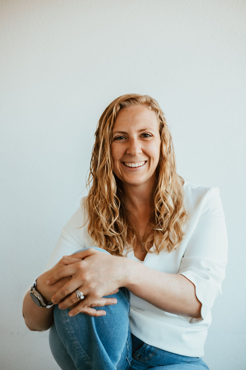 Image of Dr. Micaela Dlugosz sitting with one knee up, wearing a white shirt and blue jeans.
