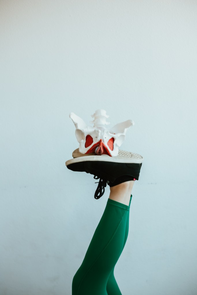 Image of a pelvic floor model being balanced on someone's feet. Person is wearing black shoes and green leggings.